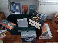 Overview of lot contents in green storage bin showing flatware sets, roasting pan with lid, boxes of utensils, cheese board, carafes, and Farberware colander box