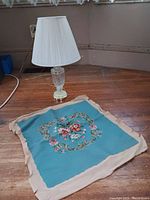Glass table lamp with white pleated lampshade and needlepoint canvas laid flat on floor.