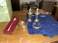 Pair of sterling silver weighted salt and pepper shakers with two sterling silver engraved cuff bracelets on blue Louis Vuitton pouch and a small red box holding a sterling iced tea spoon.