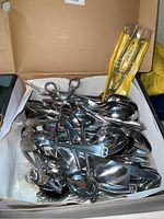 Open box displaying various metal serving spoons, some tongs, and nut crackers inside.