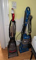 Two upright vacuum cleaners side by side against a wall, one red and black Hoover and one blue Shark, viewed from the front.