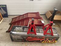 Two red MotoMaster axle stands on top of the product box with two red metal vehicle ramps behind them.