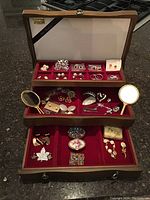 Jewelry collection inside open wooden case showing three tiers: top compartment with small slots holding earrings and pins, middle drawer with brooches, mirrors, and earrings, bottom drawer with bracelets and pill boxes.