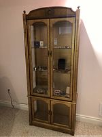 Full frontal view of a tall painted display cabinet with glass front doors showing two glass shelves inside. Lower section has two solid doors. Cabinet shows wear, located in a room with beige carpet.