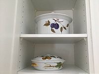 Two casserole dishes inside a cabinet shelf; one with a design of purple plums and the other with gold pears with green leaves and gold trim.