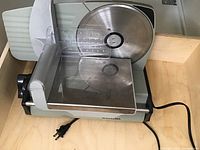 Side view of the meat slicer showing the circular stainless steel blade and sliding meat tray with plastic guard.