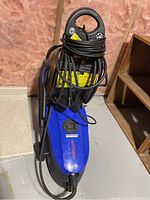 Blue Simoniz S1700 electric pressure washer with black hose and power cord, sitting on floor with wooden shelving nearby.