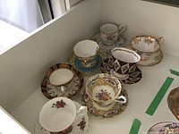 Seven different sets of Royal Albert tea cups and saucers arranged, showing variety of floral and gold designs.