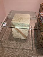 Front side view of the table showing the glass top and faux stone base on carpet floor next to patterned sofa.