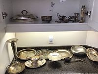 Full set of assorted silver plated and metal serving ware on dark granite countertop under cabinet light, showing covered dish, vase, gravy boat, salt and pepper shakers, candle holder, small fork tongs.