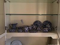 Wide view of shelf with assorted blue and white Staffordshire-style pottery including pitcher, bowls, lidded container, oval dish, and two metal blue and white plates.