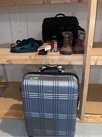 Silver hard-shell rolling suitcase by N.Y. Cargo shown with black laptop bags, boots, shoe stretcher, spray bottle, and blue gym bag on wooden shelving.