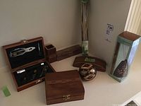 Wooden wine opener set in box with black velvet interior, assorted wooden trinket boxes in various shapes and sizes, decorative spherical ball with metallic paint, and other wood decor pieces on a floor surface.