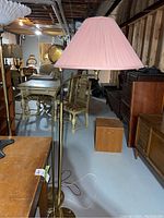 Brass floor lamp with pink pleated fabric shade illuminated from above in basement setting with other furniture around.