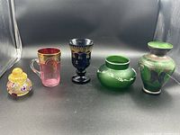 Five antique art glass pieces displayed in a row including a perfume bottle, a cup, a pedestal goblet, a small bowl vase, and a larger vase with silver accents.