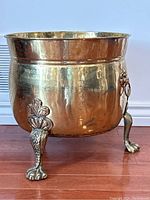 Front angled view of the brass clawfoot planter showing one claw foot and lion head ring handle