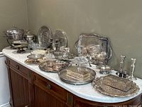 Wide view of large assortment of silver plated and stainless steel serving and decorative items on marble top surface, showing trays, bowls, candlesticks, trinket boxes, and salt shakers.