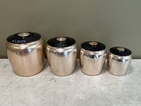 Four vintage Kromex aluminum canisters with black lids labeled FLOUR, SUGAR, COFFEE, TEA arranged by size.