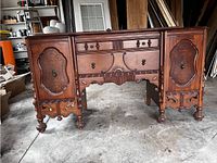 Front view of antique wooden sideboard showing two cabinet doors, three drawers, ornamental carvings and brass hardware on legs and doors.