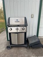 Full view of Broil King propane BBQ grill with wheels, two-door cabinet, side shelves beside it, and weathered condition visible.