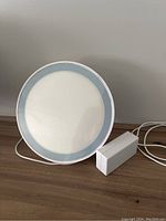 Front view of the round white light panel with pale blue trim and power adapter on a wooden surface.