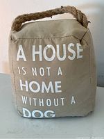 Front view of beige fabric doorstop with white text 'A HOUSE IS NOT A HOME WITHOUT A DOG' and thick braided rope handle.