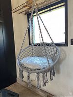 Grey woven hammock chair hanging with cushion on it, showing front and side view and decorative tassels below the seat.