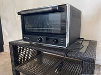 Large Kitchenaid black and silver toaster oven on woven rattan style shelf, with visible power cord and dual control knobs