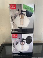 Two stacked boxes of Globe Harrow wall sconces showing the product image with clear glass shade and gold-accent socket on matte black mount.