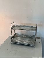 Two-tier silver metal vanity tray shown front and angled side views, highlighting lattice patterned border and ball feet.