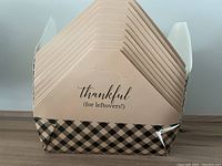 Eleven folded cardboard boxes stacked inside each other, showing the message 'thankful (for leftovers!)' on the beige flap and a black and beige plaid pattern at the bottom.
