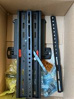 Photo showing the main metal mounting plate and articulating arm laid flat inside a cardboard box, including plastic bags of mounting hardware.
