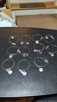 Eleven silver-tone charm bracelets displayed on a black surface. Each bracelet has small round charms with various designs and slogans. Bracelets are expandable wire bangles in typical Alex and Ani style.