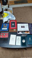 Overview of multiple coin sets displayed on a black table including a red velvet box, blue folders, coin holders, and informational pamphlets