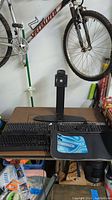 Lot of computer accessories on a table including two keyboards, mouse pad, and monitor stand with bicycle in background.