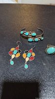 Photo of a silver-tone cuff bracelet with turquoise stones, a ring with turquoise, and a pair of earrings with multicolored stones arranged on a black surface.