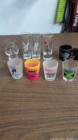 All nine assorted shot glasses on wooden table showing various printed logos and texts.