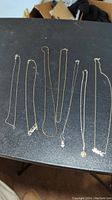 Seven silver necklaces laid out on a black surface showing various chain styles and pendants including 'LOVE' pendant and heart charm