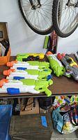 Photo showing multiple water blasters arranged on a table under hanging bike wheels. Various colors and sizes observable.