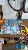 A collection of new pool toys including inflatable swim arm bands, rings, foam rings, swim noodles, and floatation devices arranged on a table under a bicycle wheel