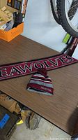 Photo of Tabor Academy knit hat with pom-pom and embroidered logo next to maroon Seawolves scarf on a wooden surface.