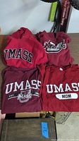 Four UMass-themed apparel items including two hoodies and two long sleeve shirts arranged on a surface.
