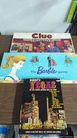 Photo showing three vintage board games stacked; Clue Classic Detective Game on top, Barbie Queen of the Prom in the middle, Weekend in Vegas at bottom.