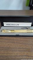 Full view of gold-tone Mini Mag-Lite flashlight in original black case with Mini Mag-Lite paper insert.