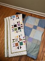 Two folded quilts side by side on a hardwood floor, showing light and dark color schemes (light quilt on left, darker quilt on right)