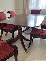 Side view of the antique double pedestal dining table showing polished wood with brass feet and caster wheels beneath the curved legs, surrounded by red upholstered chairs.