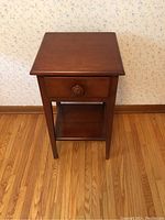 Frontal view of the solid wood bedside table showing the drawer and lower shelf, placed against a wall with wood flooring.