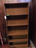 Front view of tall faux veneer pressboard bookcase with five shelves, showing the uniform medium brown wood grain finish and straight sides.