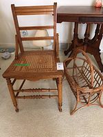 Wooden antique chair with cane woven seat alongside rattan magazine rack on carpeted floor with side table in background