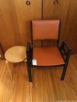 Photo showing the Mid Century style orange vinyl upholstered arm chair with dark wooden frame, alongside a round plywood side table, all placed on wooden flooring against wood paneled wall.